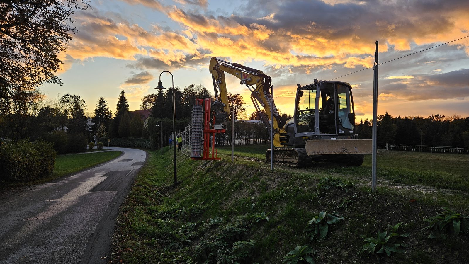 Bagger mit Drahtrollen-Abroller beim Zaunbau vor dramatischem Abendhimmel